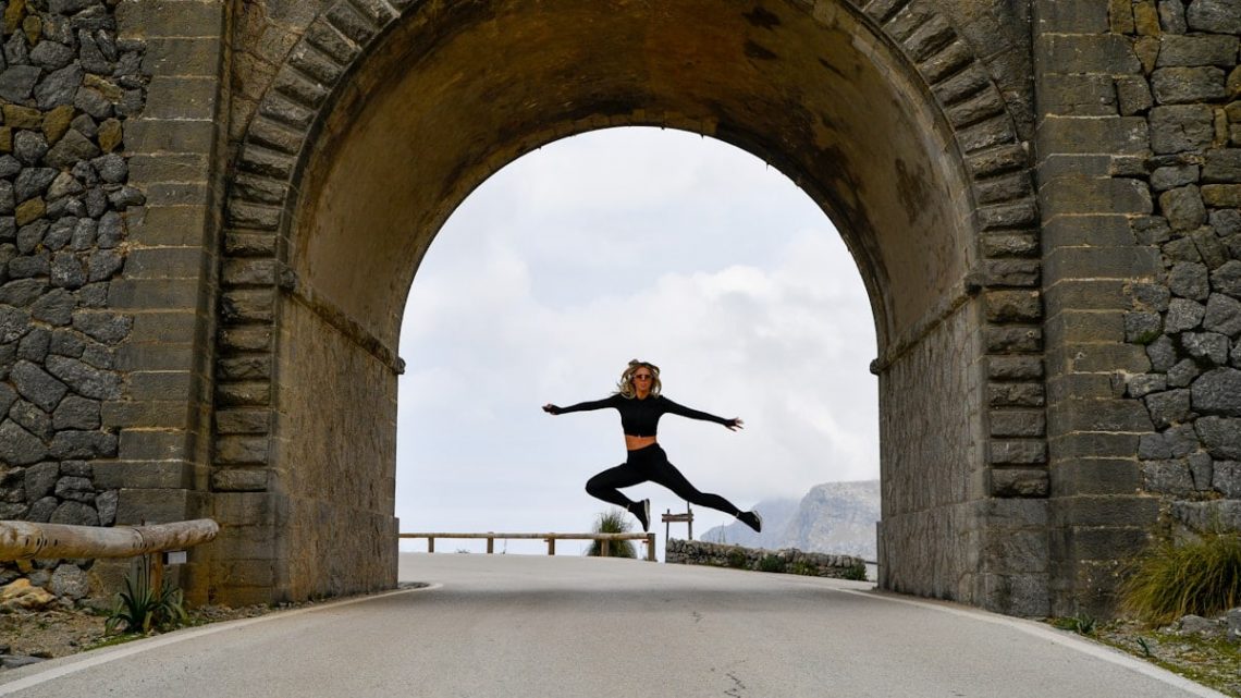 Dynamic wide-shot of an athletic young woman jumping high in the air, framed perfectly by a massive historic stone archway on a scenic mountain road. 📸 Photo by me IG: @margoevardson
