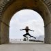 Dynamic wide-shot of an athletic young woman jumping high in the air, framed perfectly by a massive historic stone archway on a scenic mountain road. 📸 Photo by me IG: @margoevardson